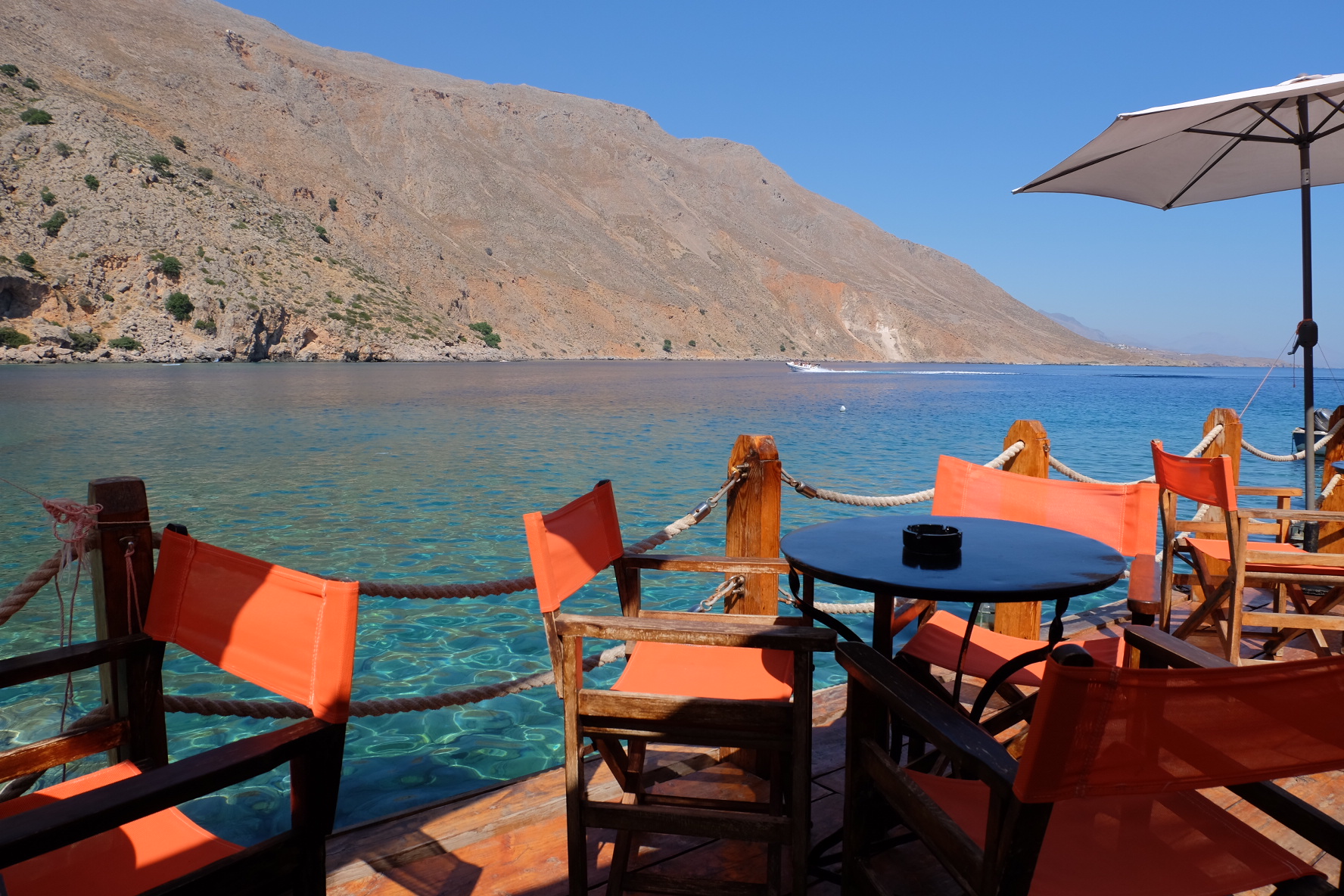 Seaside cafe with orange chairs and a table, overlooking clear blue water and rocky hills under a clear blue sky.